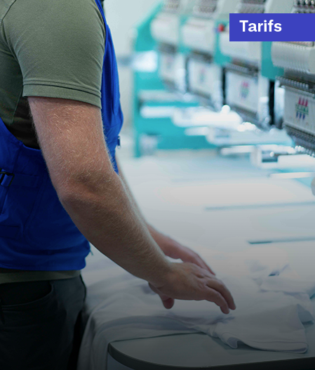 Un homme travaillant sur une machine à broder industrielle, avec des textiles blancs en premier plan, illustrant les tarifs de personnalisation ou de production textile.