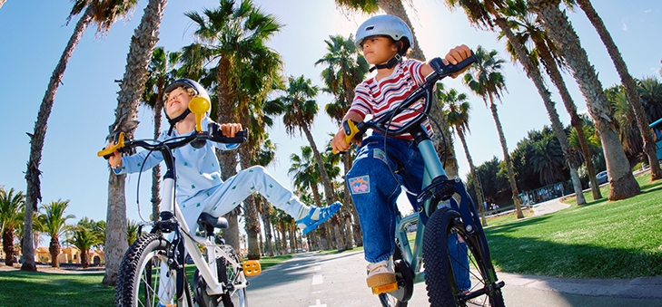 Enfants joyeux à vélo, souriant et s'amusant sur un chemin ensoleillé bordé de palmiers.